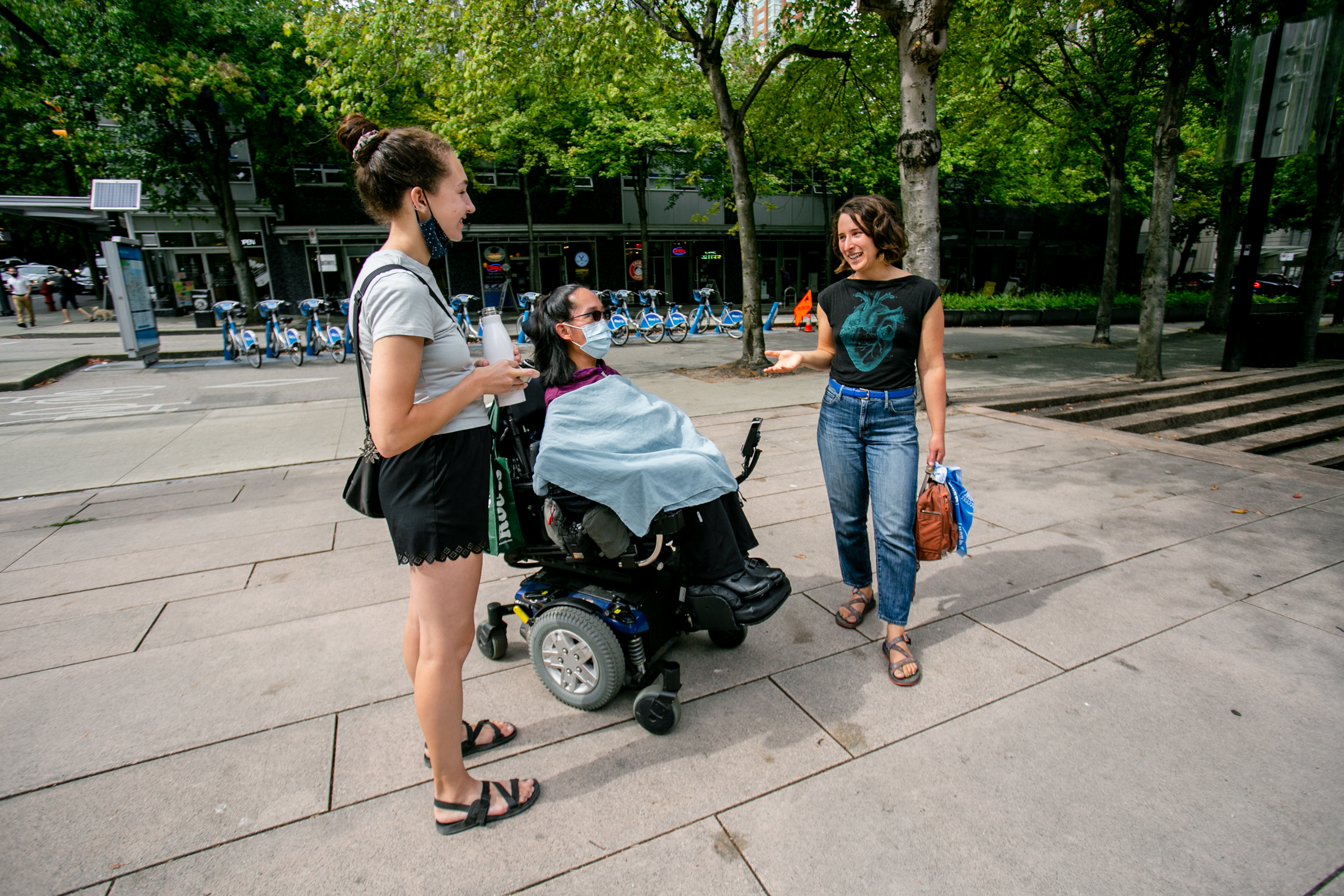 a woman using a power wheelchair and two standing people smile mid conversation. behind them is a series of bike shares.