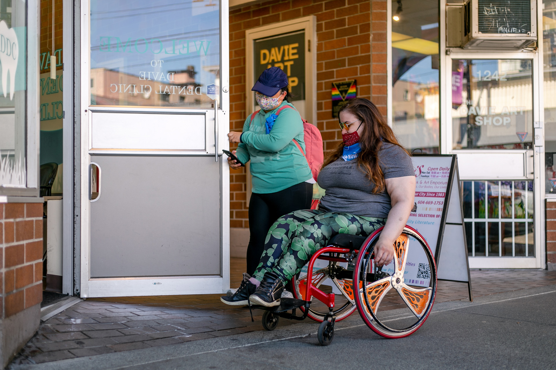 a woman in a powerwheelchair with cool orange rims and colourful leggings enters through a door being held open by someone standing