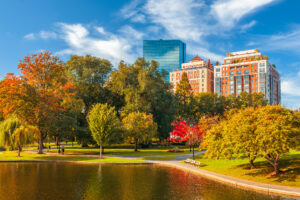 A picturesque view of Boston, Massachusetts during autumn, showcasing vibrant fall foliage with trees displaying shades of orange, red, and green. A calm pond reflects the bright blue sky, and tall buildings with glass facades and brick exteriors rise in the background. A pathway winds through the park, with benches and a few people strolling in the peaceful scene.