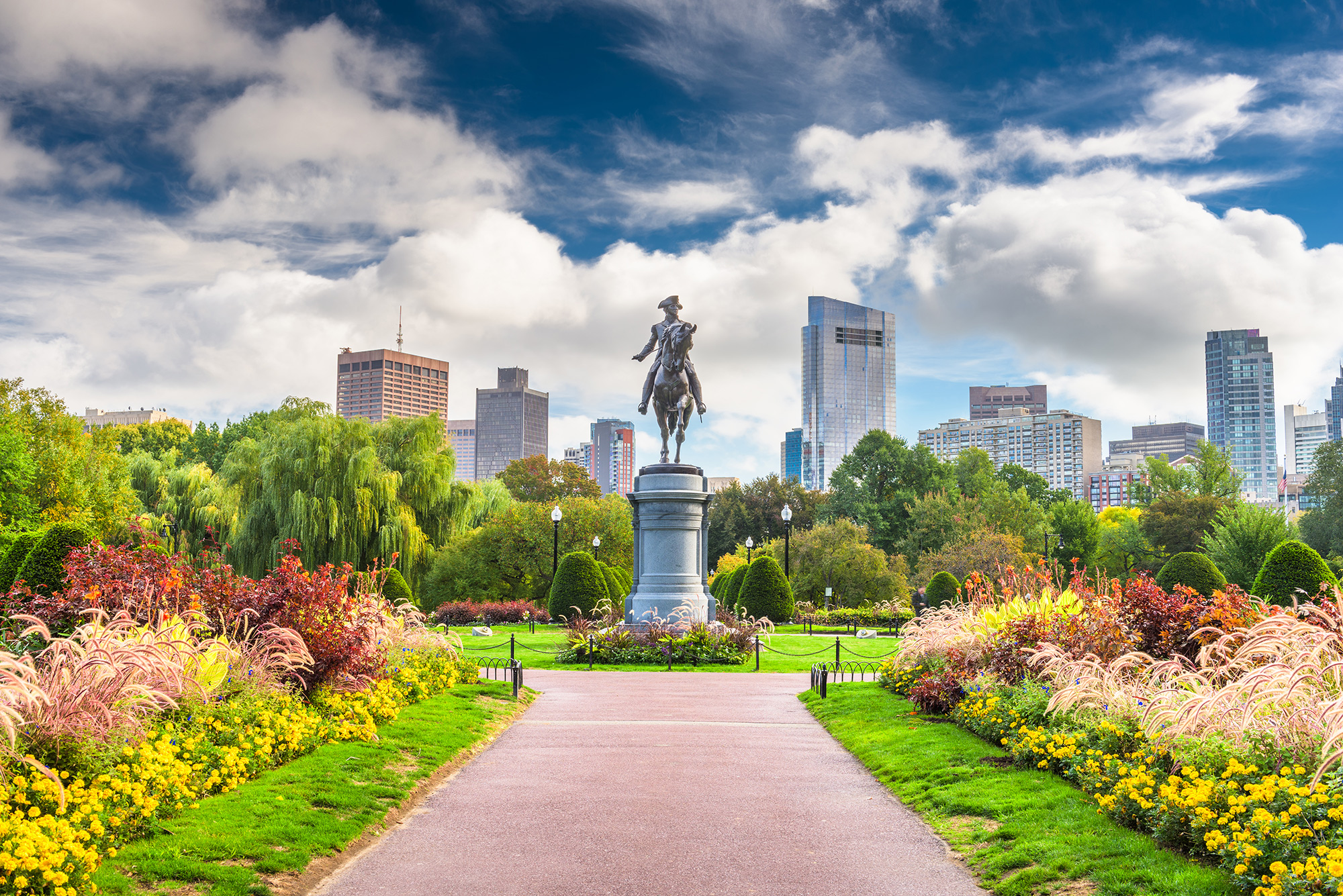 A view of the equestrian statue of George Washington in the Boston Public Garden, surrounded by colorful flower beds and well-manicured lawns. Tall modern buildings rise in the background, while the lush greenery of trees fills the park. The bright sky with scattered clouds enhances the beauty of the scene.