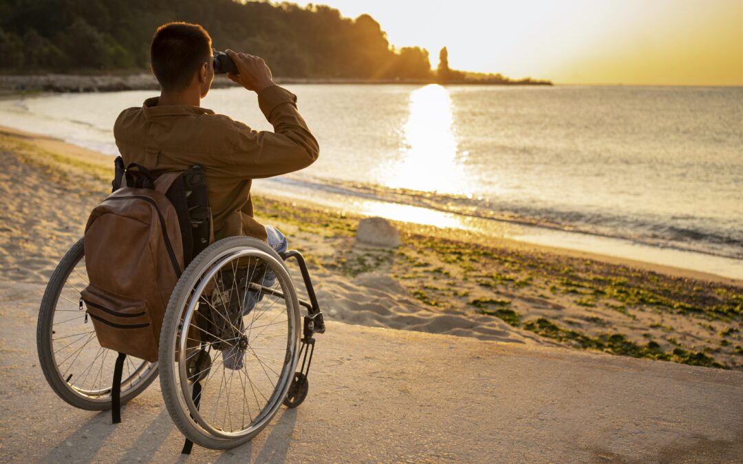 A man is seated in a wheelchair and he is facing the sunset. He is on a sandy beach, and is using binoculars to watch the orange-yellow sunset.