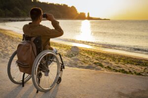 A man is seated in a wheelchair and he is facing the sunset. He is on a sandy beach, and is using binoculars to watch the orange-yellow sunset.