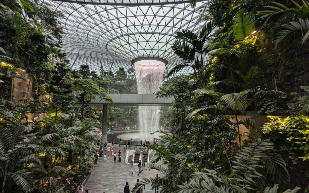 Inside Jewel Changi Airport, Singapore. A lush waterfall view. The building has a beautiful dome, surrounded by greenery and yellow lighting. In the middle is a waterfall and a bridge.