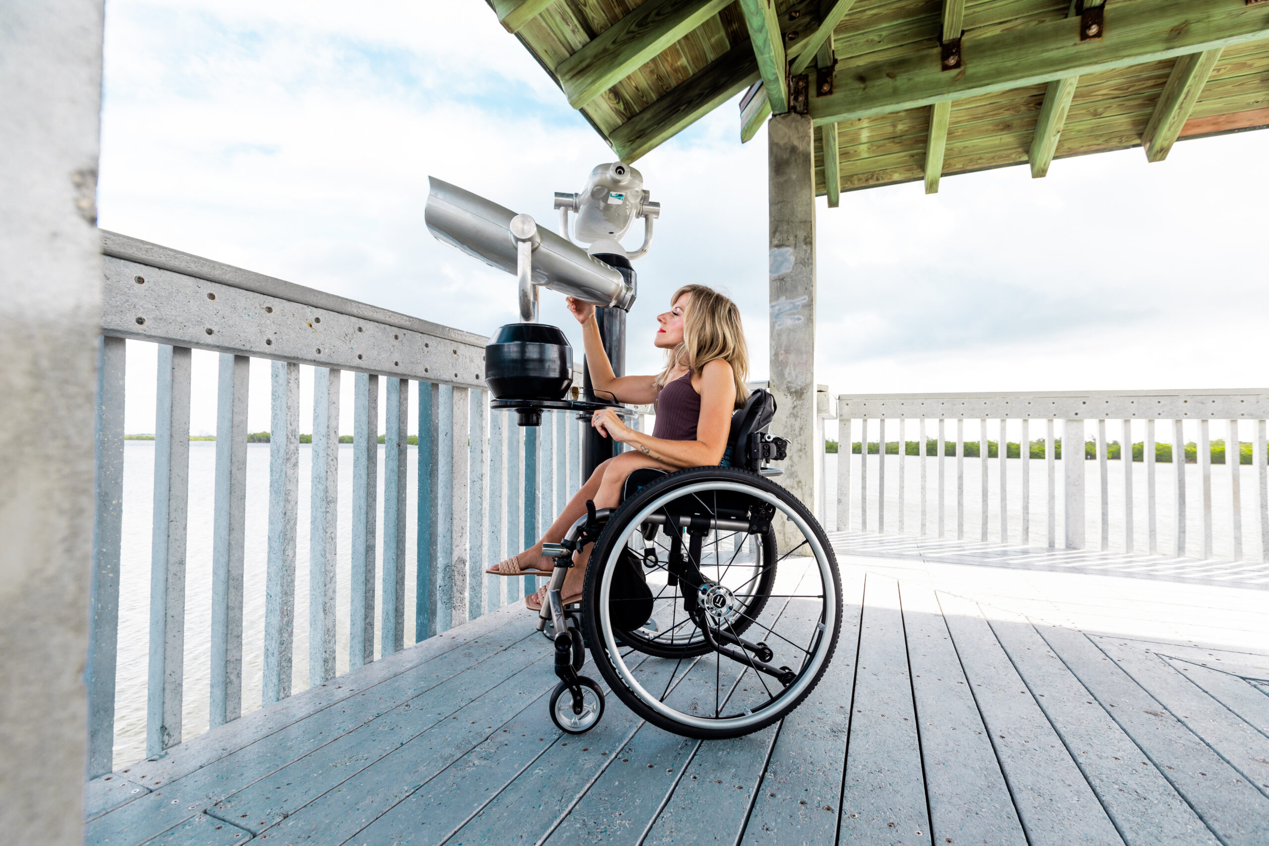 Kristin is looking out some binoculars that are perfect for her height on her wheelchair. She is at Anne Klob Nature park, wearing a brown top and a beige flats.