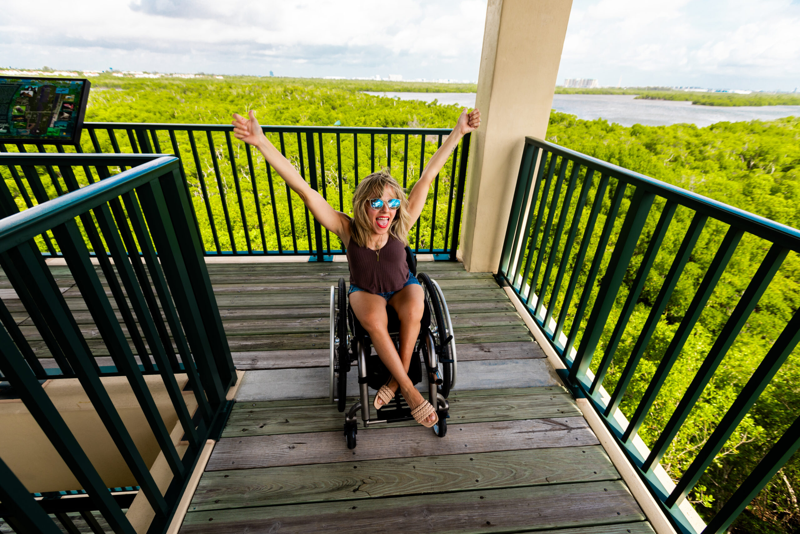 Kristin is at the top of an observation tower at Anne Klob Nature park in Fort Lauderdale. She is wearing sunglasses and a brown top. Her hands are in the air and she is smiling. Behind her is beautiful green scenery.