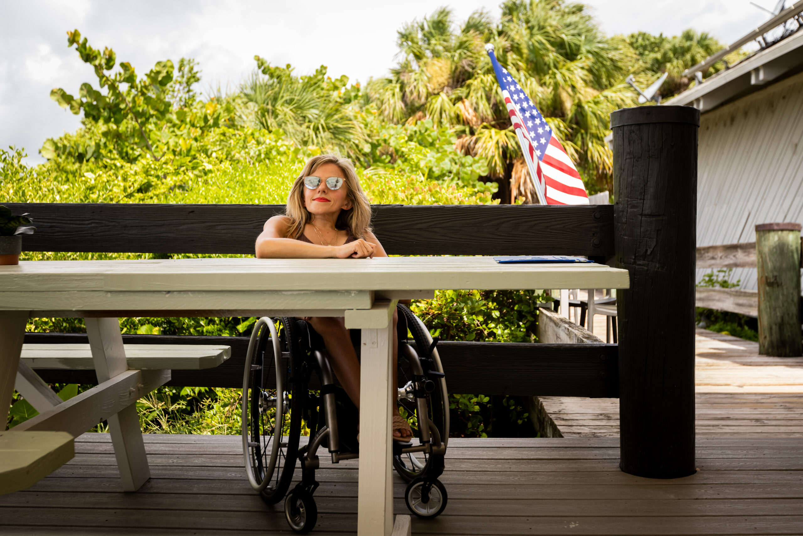 Kristin is on a rooftop, she's seated in her wheelchair, wearing sunglasses. Her arms are resting on a table. She is smiling, and the American flag is waving behind her.