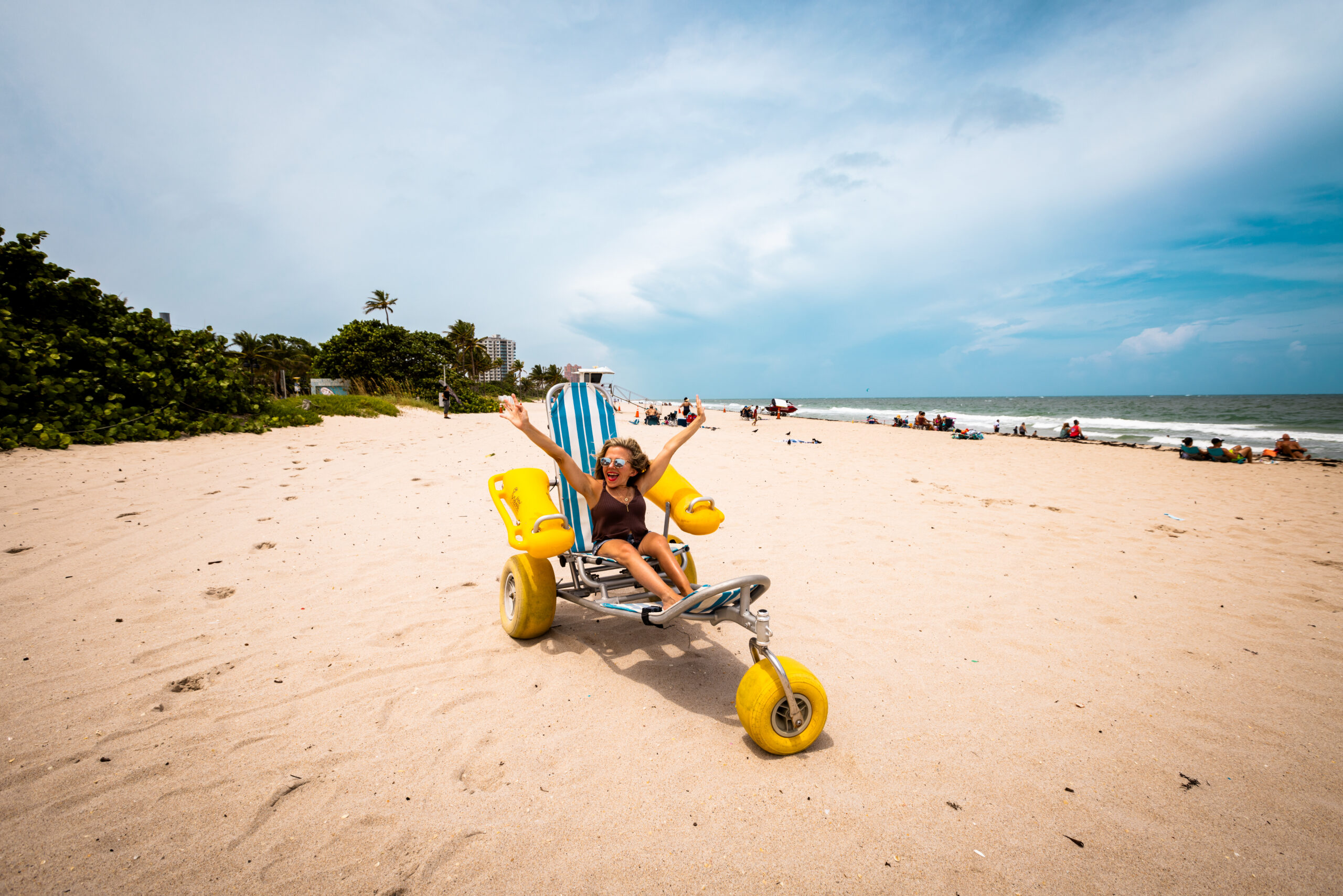 Kristin is seated on a blue, yellow and white beach wheelchair at Hugh Taylor Birch State Park. She is surrounded by sand, and in the distance are other people and the ocean.