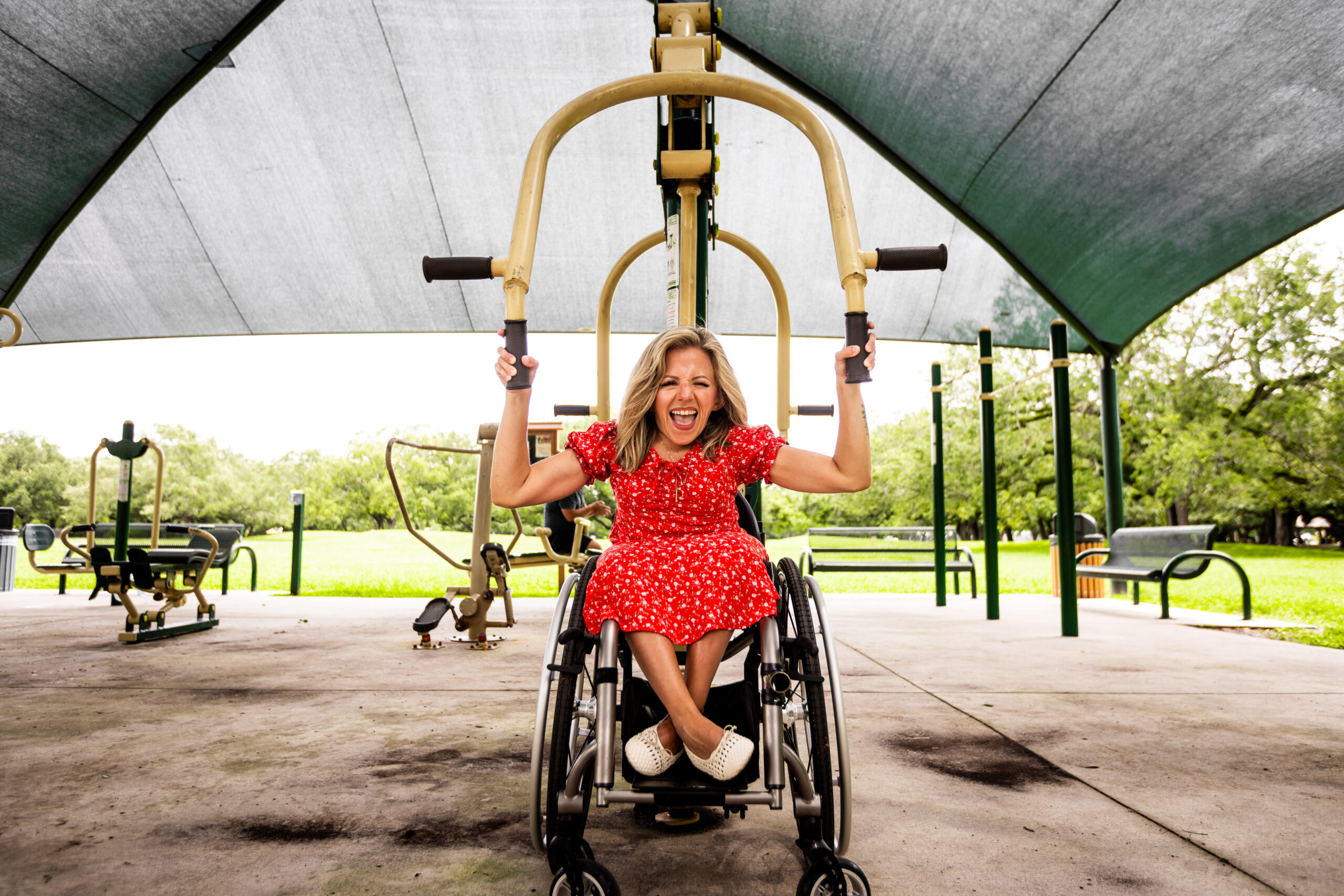 Kristin at the Topeekeegee Yugnee (TY) Park, exploring their accessible fitness zone. She is wearing a a red dress with white flowers on it, smiling and winking at the camera.