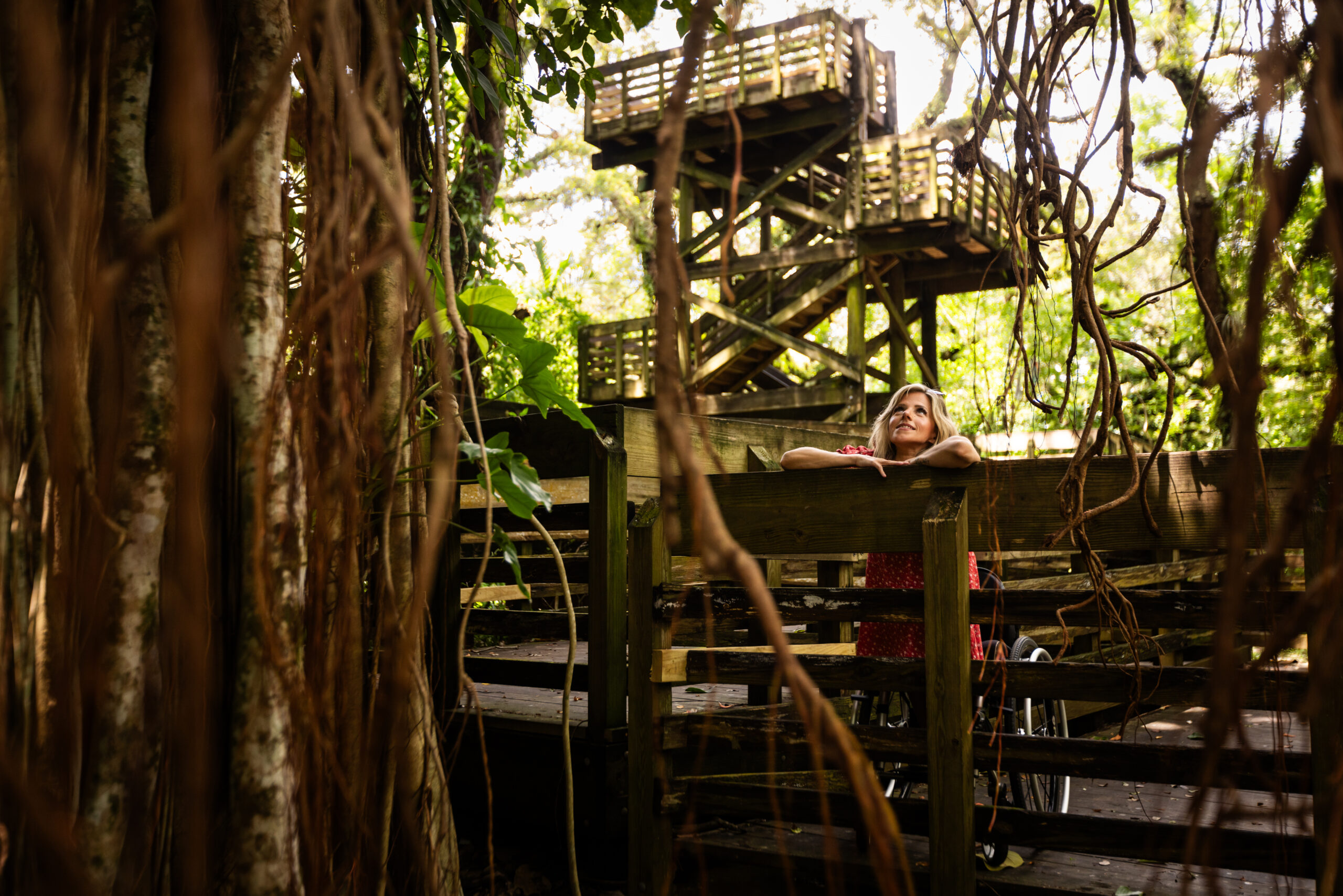 Kristin is in the middle of a beautiful, woody trail at Tree Tops Park. She is seated on her wheelchair, wearing a red and white dress, her arms are resting on some wood as she looks up at the sky.