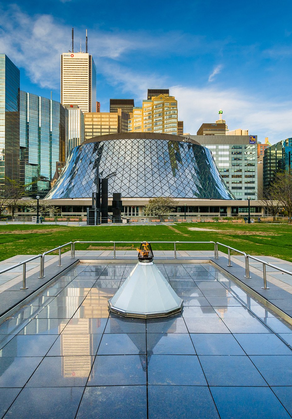 View of David Pecaut Square in downtown Toronto, surrounded by modern skyscrapers under a clear blue sky. In the foreground is a polished stone plaza with stainless steel railings and a central glass structure housing the Eternal Flame, which burns brightly.