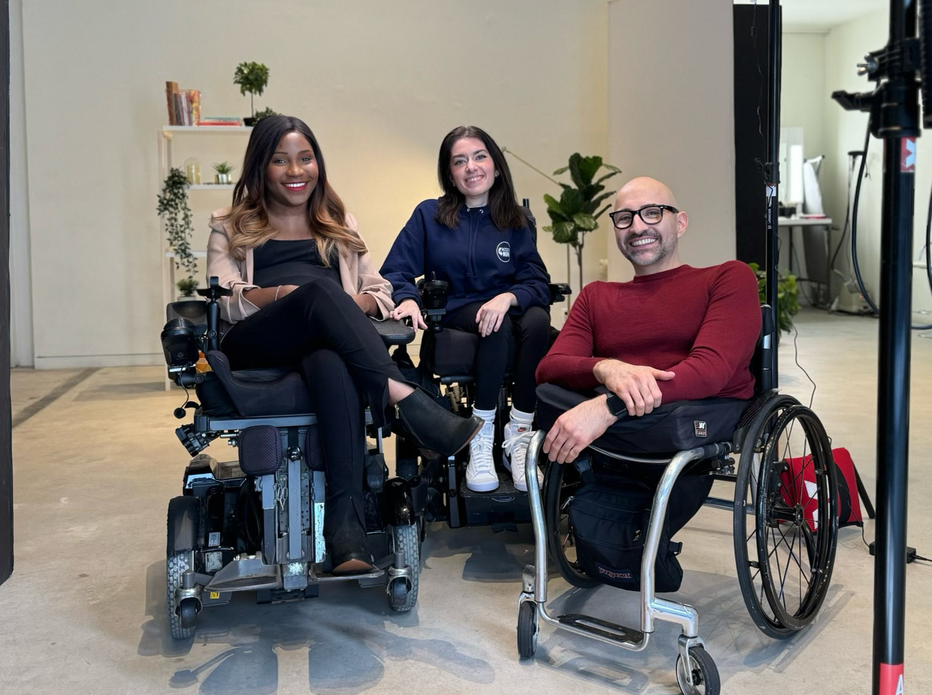 IMG_6128 Three disabled advocates sit smiling in a bright studio space. Two people use power wheelchairs and one uses a manual wheelchair. The woman on the left wears a blush blazer and black outfit, the woman in the middle wears a navy hoodie and sneakers, and the man on the right wears glasses and a red sweater. They are surrounded by plants and studio equipment, creating a professional yet relaxed atmosphere.