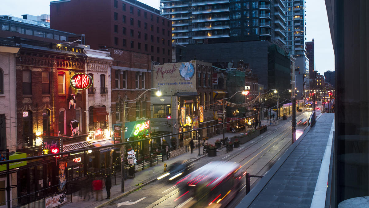 King_Street_West_night_7c23d2c5-2c5c-4b3b-a0d5-938c2fac0200 Evening view of King Street West in Toronto, showing a vibrant row of restaurants and bars with neon signs, historic brick buildings, and a blur of streetcar lights in motion, with high-rise condos in the background.