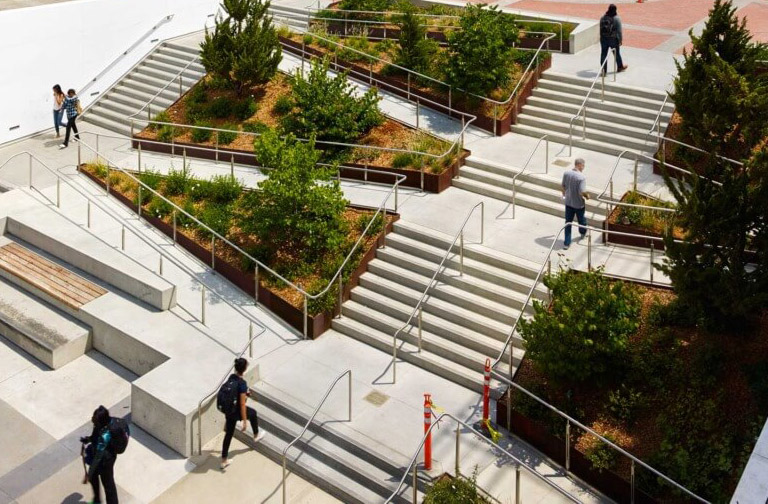 clay-holden-ucb-lower-sproul-24-1024x580 A modern, multi-level outdoor plaza featuring a series of wide staircases and switchback ramps surrounded by lush greenery. The design integrates accessibility with aesthetic landscaping, offering handrails and resting areas throughout. Several people are walking along the paths and stairs.