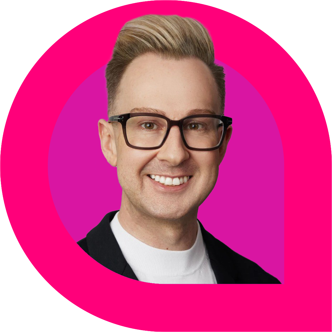 a person with styled blonde hair and dark-rimmed glasses, smiles brightly while wearing a white top and black blazer