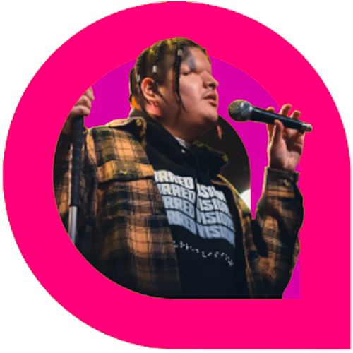 Mattmac, an artist with braided hair, holds a microphone while performing. He wears a plaid jacket over a hoodie with bold text, framed within a vivid pink graphic shape. The stage lighting casts a warm glow as he appears mid-performance.
