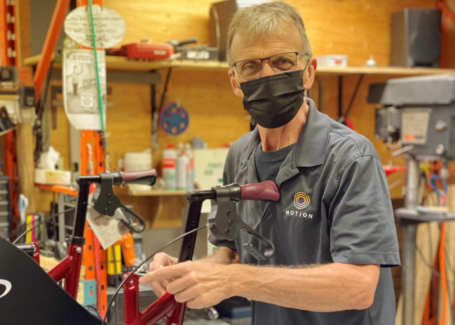 A technician wearing a black face mask and glasses works on a mobility device in a workshop. He wears a dark grey polo shirt with the logo for Motion, a company specializing in mobility and accessibility solutions. The background includes tools, shelves, and workshop equipment.