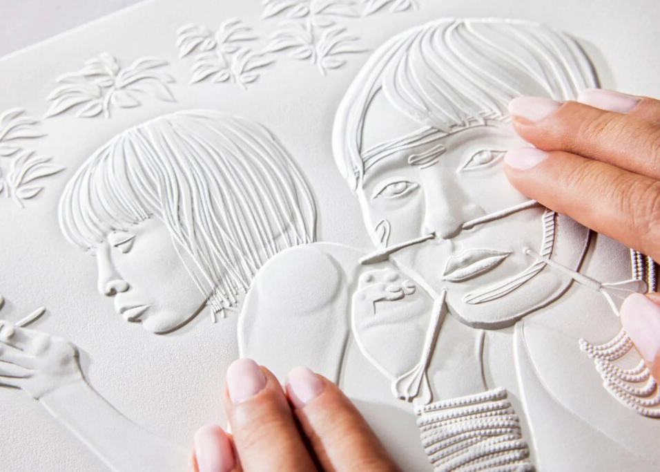 A close-up of hands exploring a tactile artwork featuring two people’s faces and detailed elements like hair, jewelry, and plants. The image is embossed on white paper, allowing blind or low vision individuals to feel the contours and textures of the visual scene.