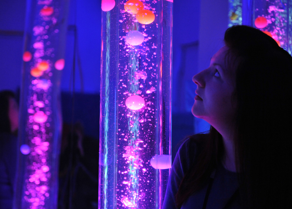 A young woman gazes at a tall, cylindrical bubble tube filled with glowing, floating orbs illuminated in vibrant shades of pink, purple, and orange. The surrounding space is dimly lit with a soothing blue hue, creating a calming, sensory-rich environment. Similar bubble tubes can be seen in the background, adding to the immersive experience.