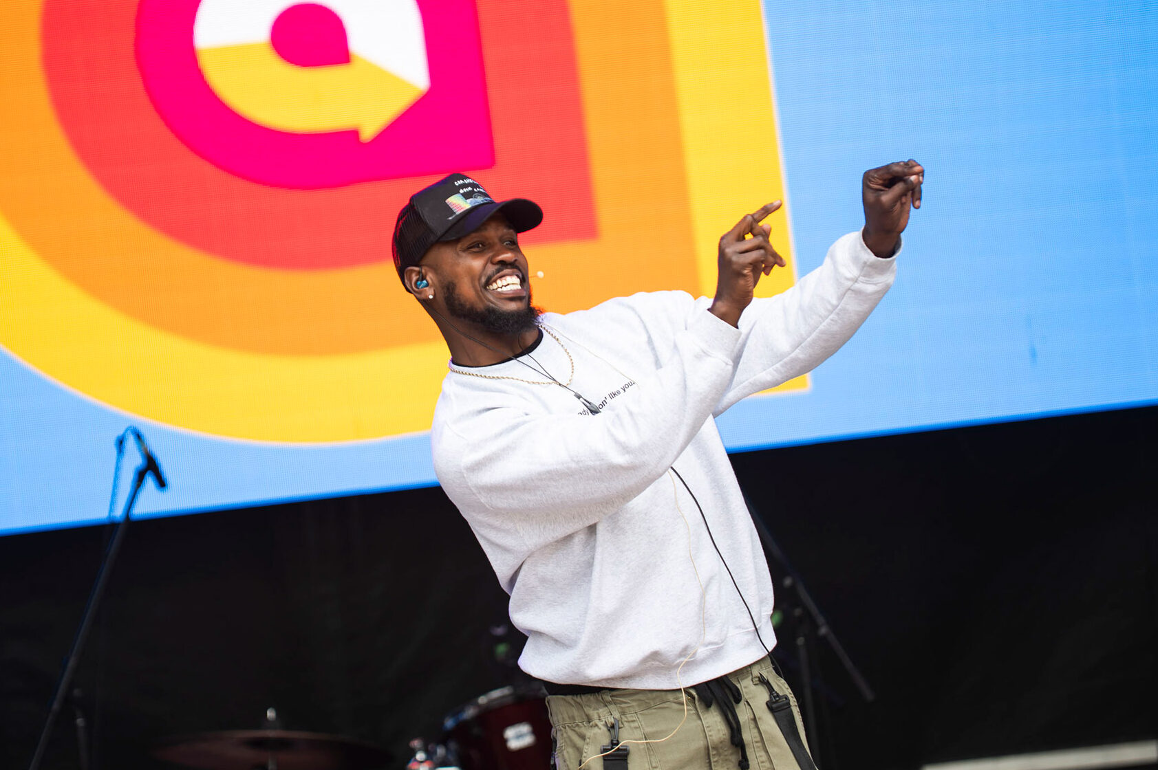 Performer on the AccessFest main stage mid-dance, smiling energetically with arms raised. He wears a grey sweatshirt, olive cargo pants, and a black cap. Behind him is a vibrant screen featuring the AccessFest logo in red, orange, and yellow concentric shapes