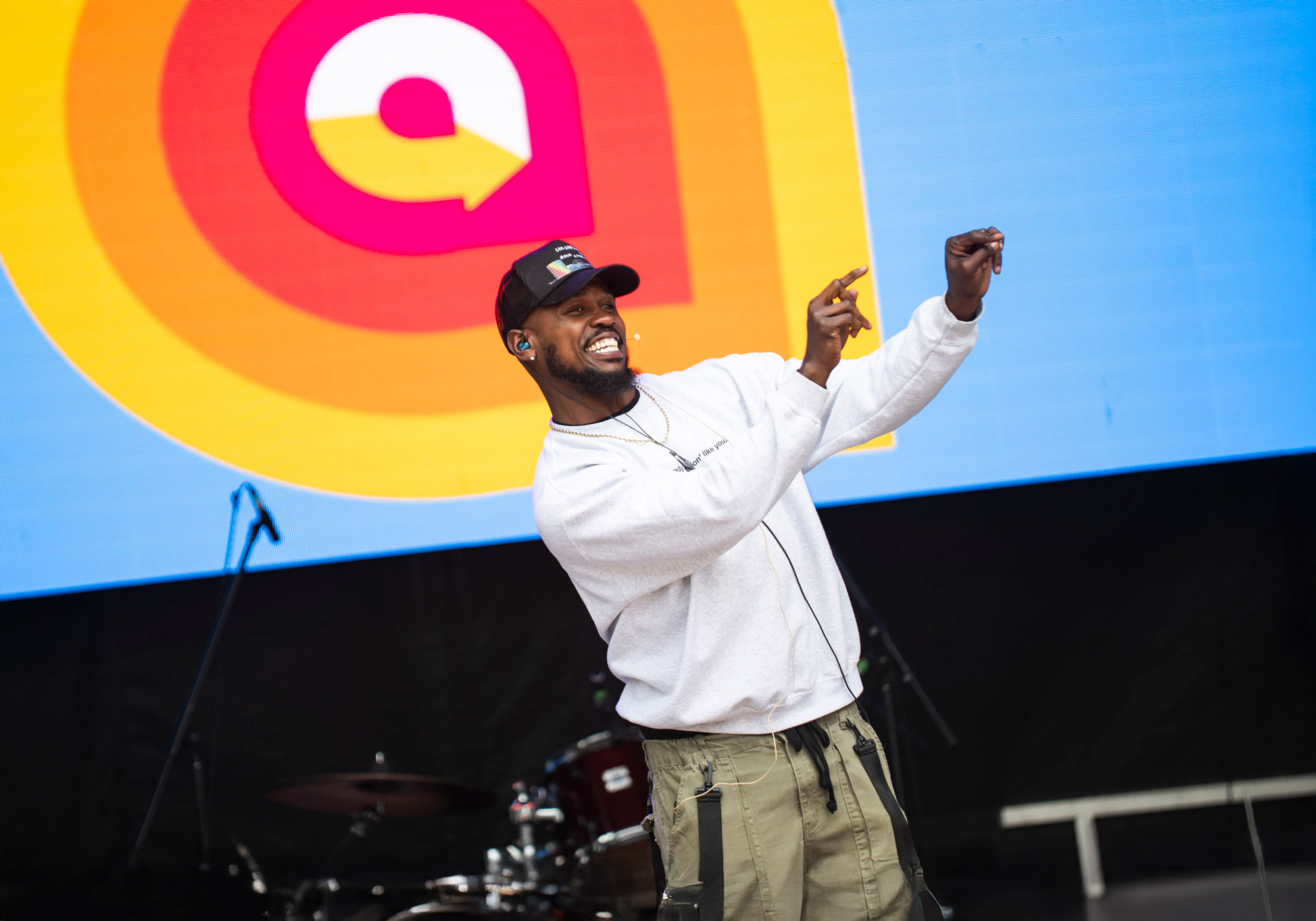 Performer on the AccessFest main stage mid-dance, smiling energetically with arms raised. He wears a grey sweatshirt, olive cargo pants, and a black cap. Behind him is a vibrant screen featuring the AccessFest logo in red, orange, and yellow concentric shapes