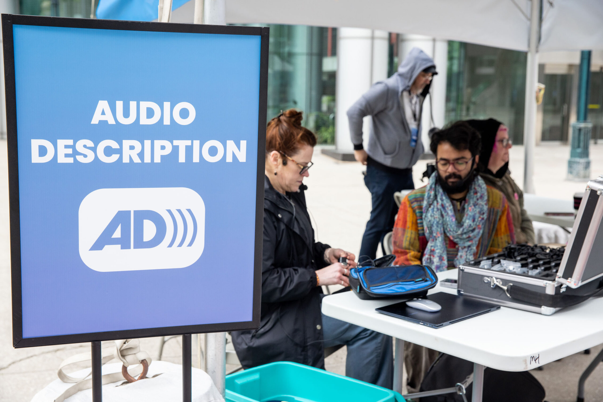 Accessfest25_WEP_05.31.25-163 A blue sign reads “Audio Description” in bold white letters, with a white icon featuring the letters “AD” followed by three curved lines indicating sound. The sign is next to a table holding tech equipment, including a laptop and cases, under a canopy in an urban outdoor setting.