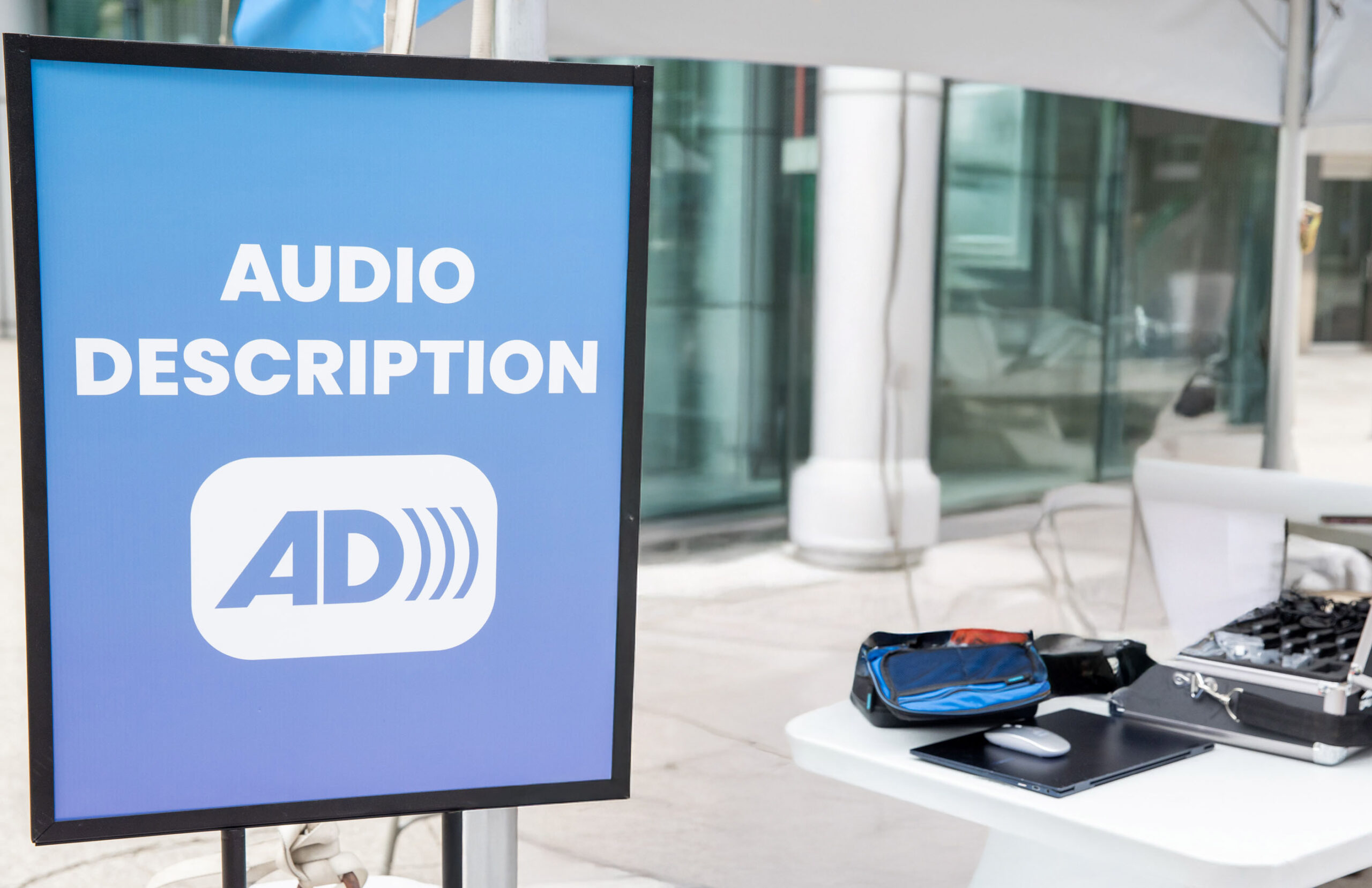A blue sign reads “Audio Description” in bold white letters, with a white icon featuring the letters “AD” followed by three curved lines indicating sound. The sign is next to a table holding tech equipment, including a laptop and cases, under a canopy in an urban outdoor setting.