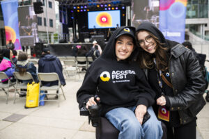 Two smiling women pose together in front of an outdoor stage at AccessFest. One woman sits in a power wheelchair wearing a black hoodie that reads “ACCESS FEST by AccessNow,” while the other leans in close, wearing a black leather jacket and hoodie with an event lanyard. Rows of seated attendees and a large screen with the AccessNow logo are visible behind them.