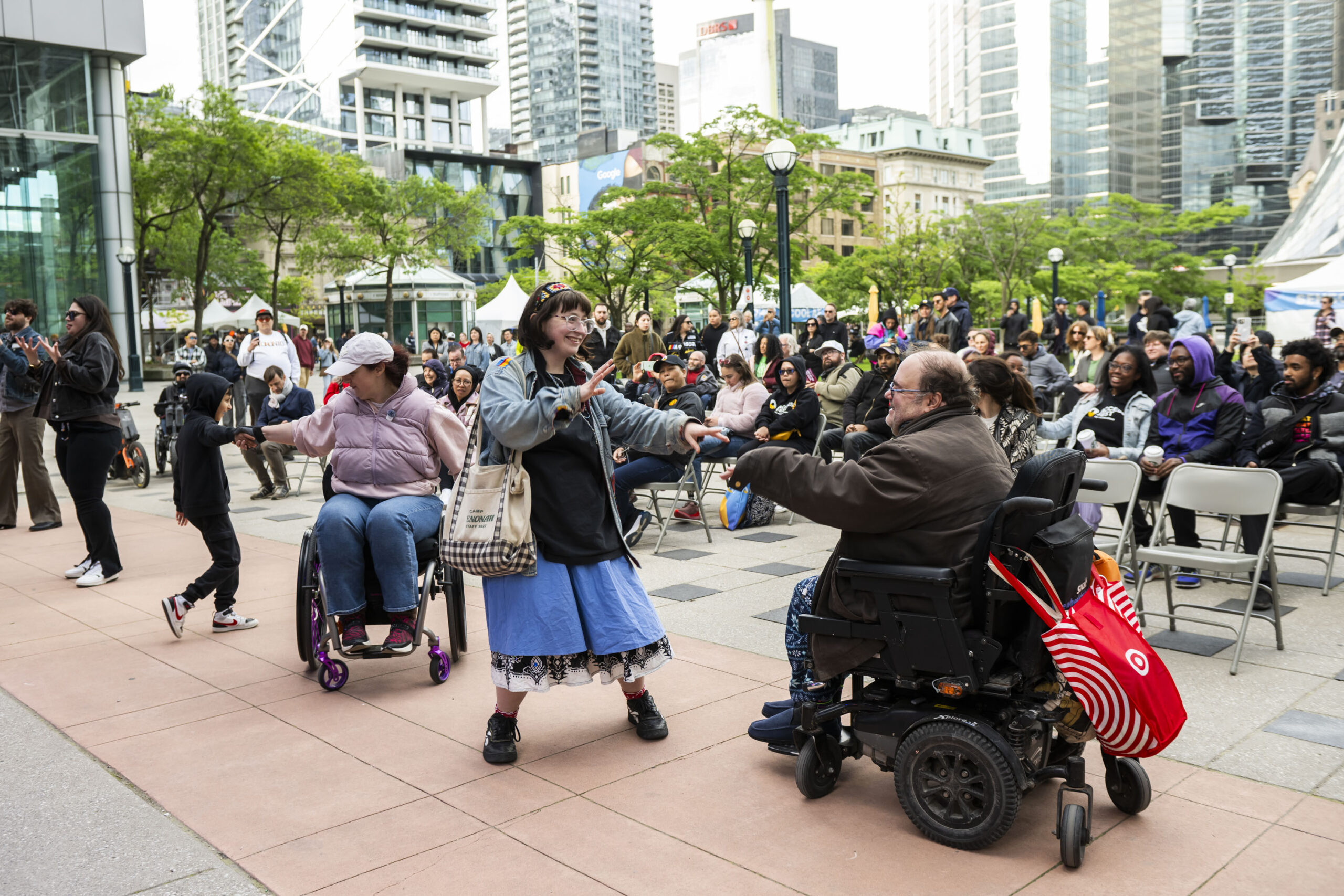 A lively crowd gathers in an urban plaza surrounded by tall buildings, with people clapping and dancing. In the foreground, three people—two using wheelchairs and one standing—joyfully dance together. Behind them, audience members seated in folding chairs watch and smile. The scene captures a vibrant, inclusive celebration in a public outdoor space.