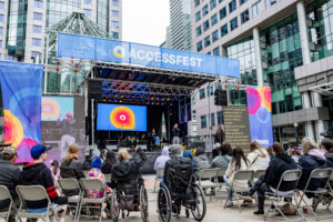 AccessFest stage is lit up with Maayan Ziv on the stage with an ASL interpreter. She is surrounded by a crowd of people seated in chairs or wheelchairs.