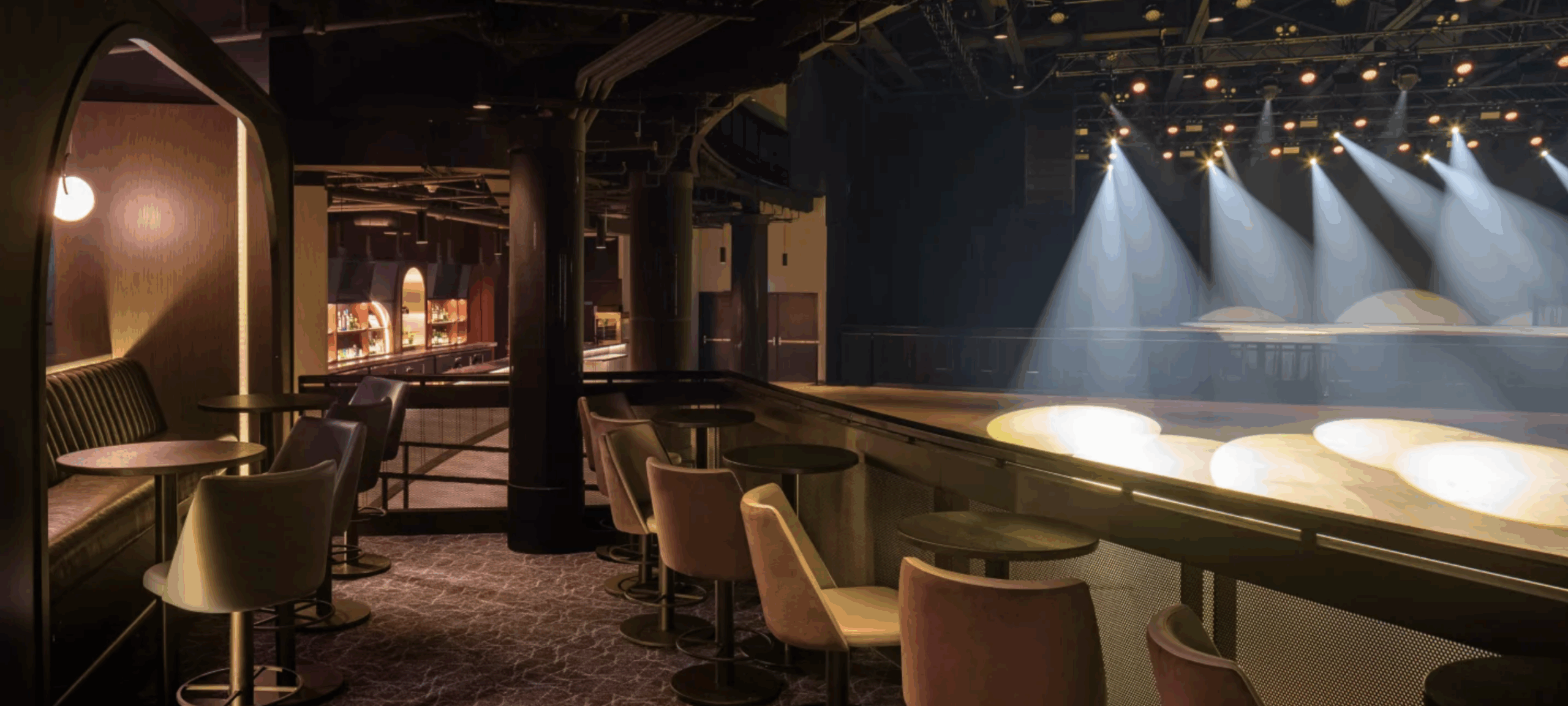 history A modern concert venue with moody lighting and an upscale lounge-style balcony. The image shows intimate seating areas with round tables and upholstered chairs overlooking a stage lit by beams of white spotlights. A sleek bar with illuminated shelves is visible in the background. The atmosphere is stylish and sophisticated, designed for a comfortable viewing experience
