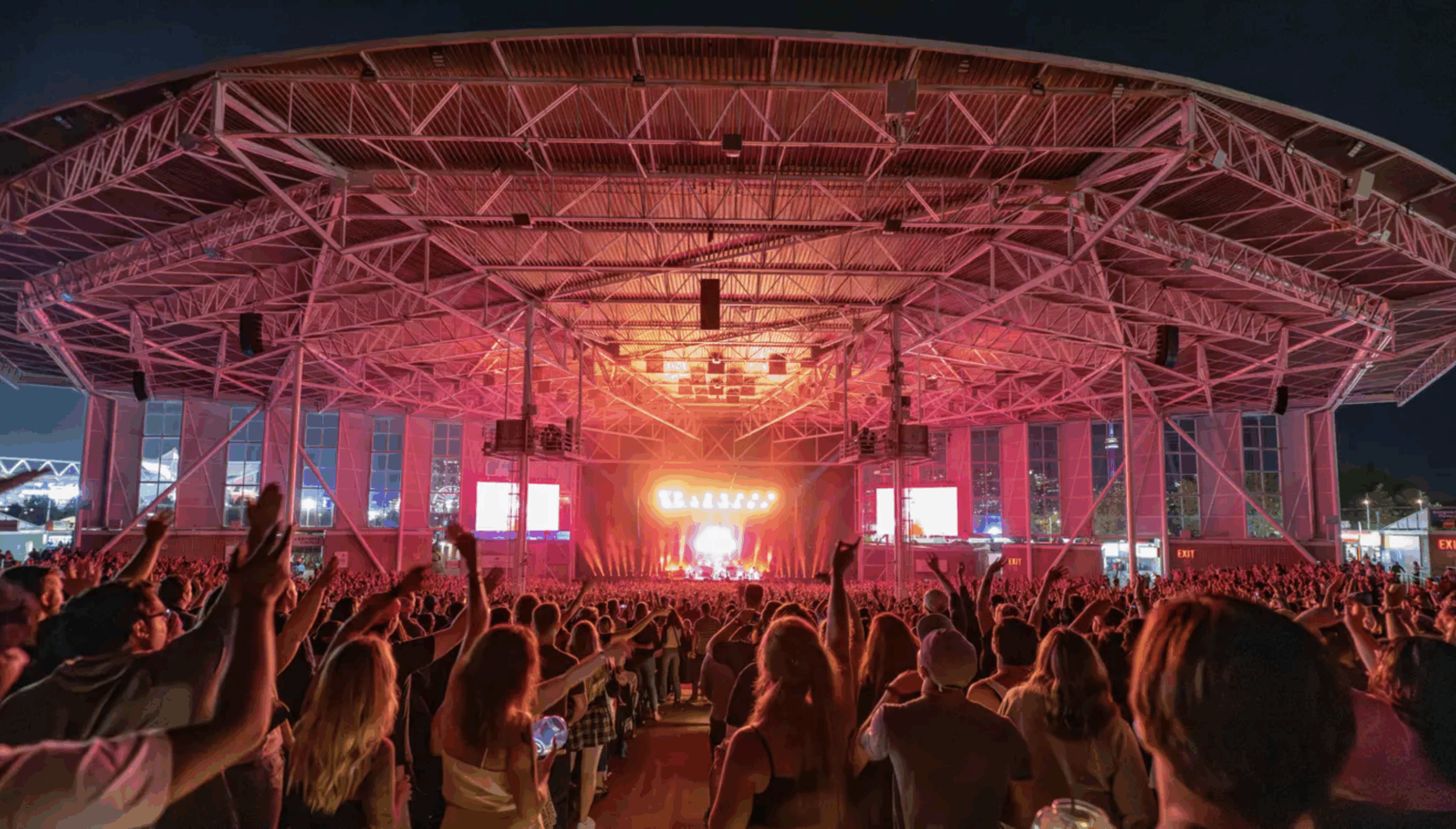 budweiser A large crowd watches a concert under the expansive open-air roof of History Toronto. The audience is silhouetted against the bright stage lights, which glow orange and red, casting dramatic beams upward. The venue's steel roof structure and support beams are illuminated, with exit signs visible along the walls. The energy is electric as hands are raised and people dance to the music.