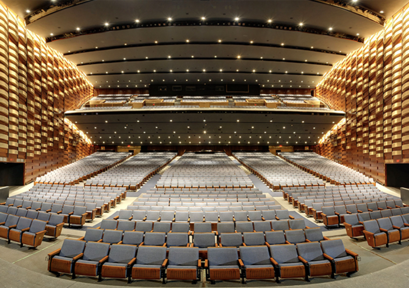 meridian hall The space features wide aisles and neatly aligned rows of grey cushioned seats with wooden armrests. The venue has multiple tiers, including a balcony and mezzanine, and distinctive curved wooden paneling along the walls. The atmosphere is grand and contemporary, with well-distributed overhead lighting creating a warm and inviting ambiance.