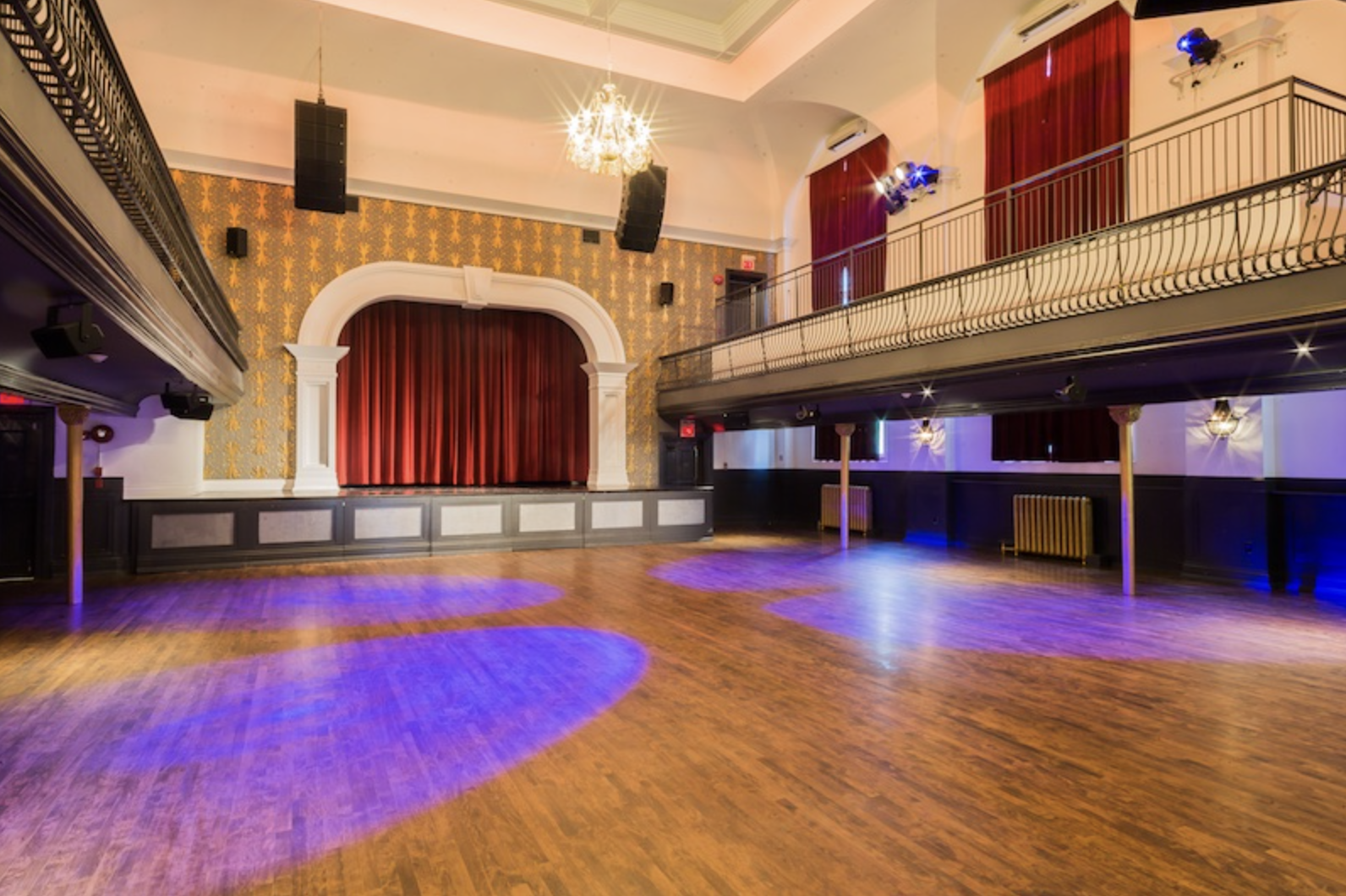 great hall Interior of a vintage-style performance venue with an elegant wooden floor, a raised stage framed by white pillars and red velvet curtains, and a decorative golden wallpapered backdrop. The space is empty and lit with purple and blue accent lights. A balcony with black railings wraps around the upper level, and chandeliers hang from the high, white ceiling. The venue has a mix of classic and modern features, and the main floor appears step-free.