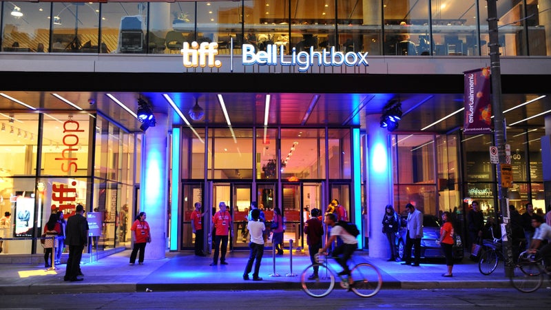 tiff bell lightbox Exterior view of the TIFF Bell Lightbox in downtown Toronto at night. The modern glass building is brightly lit with blue and purple lights, and people are gathered outside, some in red volunteer shirts. The entrance is accessible with automatic doors and a wide, step-free pathway. A cyclist rides past in the foreground, while others walk or stand nearby, creating a lively, urban atmosphere. The TIFF shop is visible through the glass on the left.