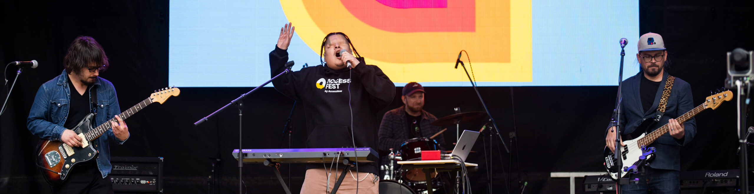 A live performance at AccessFest with Mattmac singing into a microphone at center stage, one hand raised. He wears a black AccessFest hoodie and beige pants, standing in front of a keyboard. Behind him is a large digital screen displaying the AccessFest logo. Band members play electric guitar, bass, and drums on either side of him.