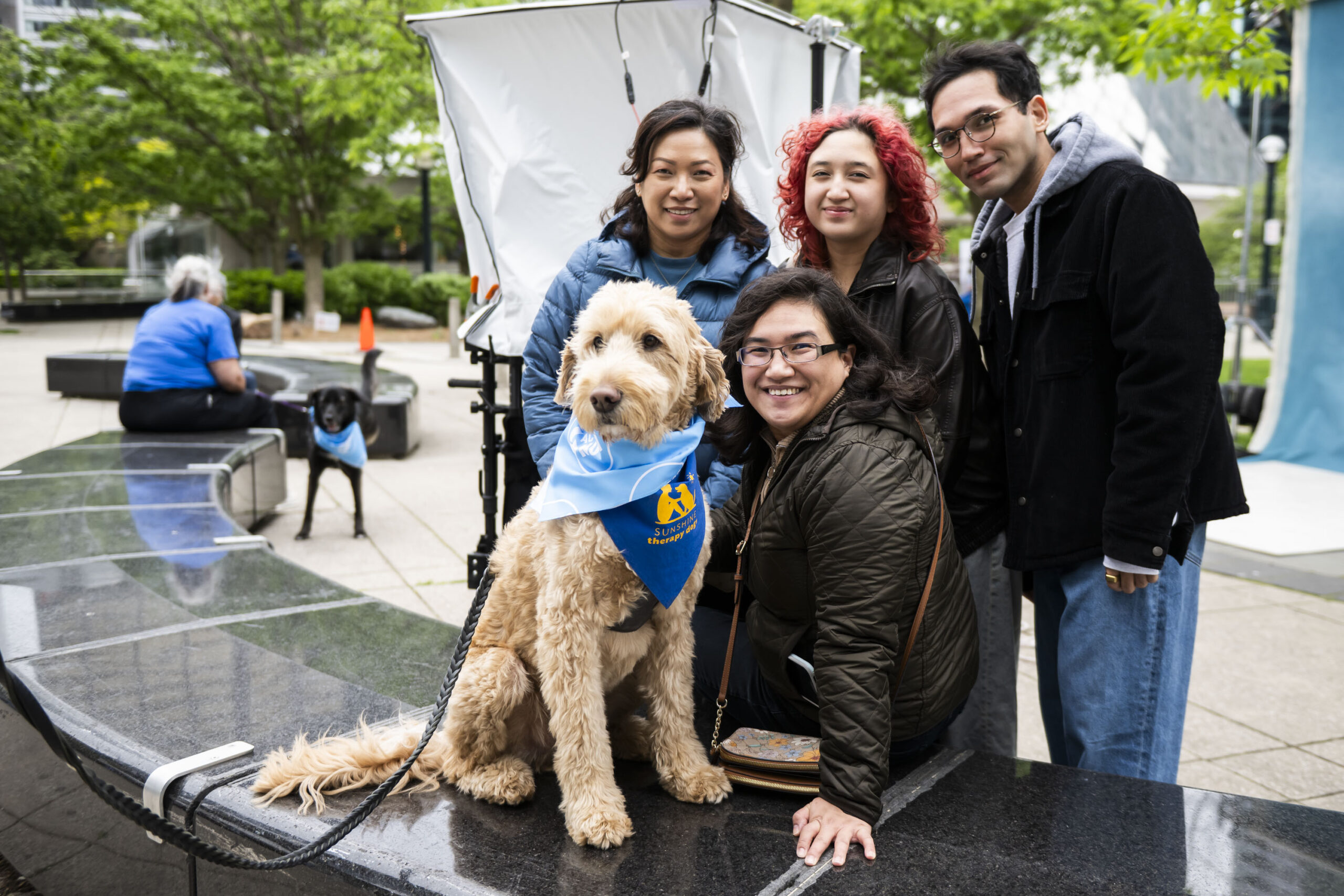 Four people smile while posing with a large golden doodle therapy dog wearing a blue bandana that says “Sunshine Therapy Dogs.”