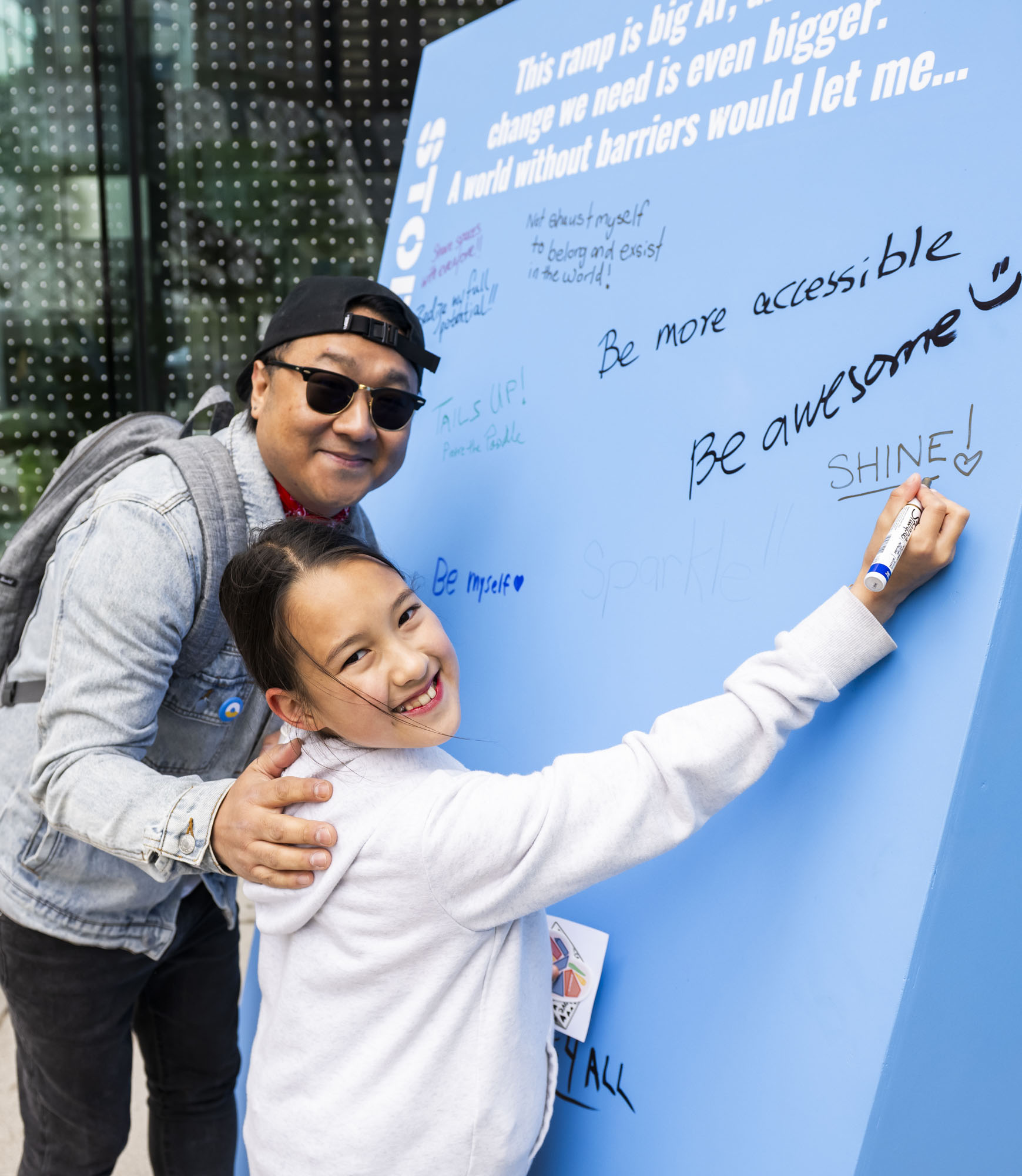 A young girl smiles while writing on a large blue ramp-shaped wall installation with phrases like “Be more accessible,” “Be awesome,” and “Be myself.” She is accompanied by her father, who wears sunglasses, a backwards cap, and a denim jacket, with one arm around her shoulders. The installation reads, “This ramp is big. The change we need is even bigger. A world without barriers would let me…”