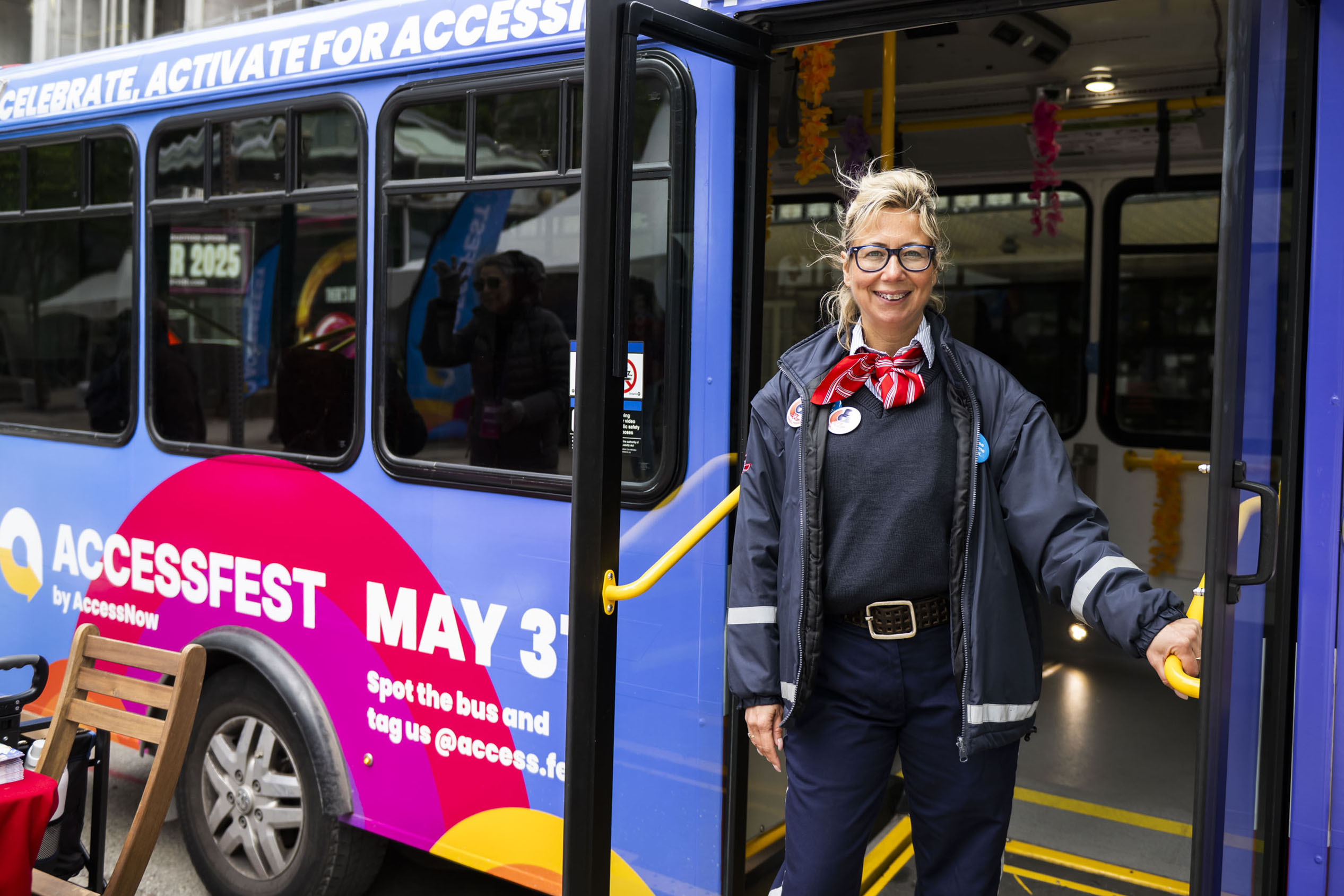 A smiling transit operator stands at the open door of a brightly wrapped AccessFest shuttle bus. She wears a navy uniform with a red-and-white striped scarf and round glasses, framed by a light ponytail.