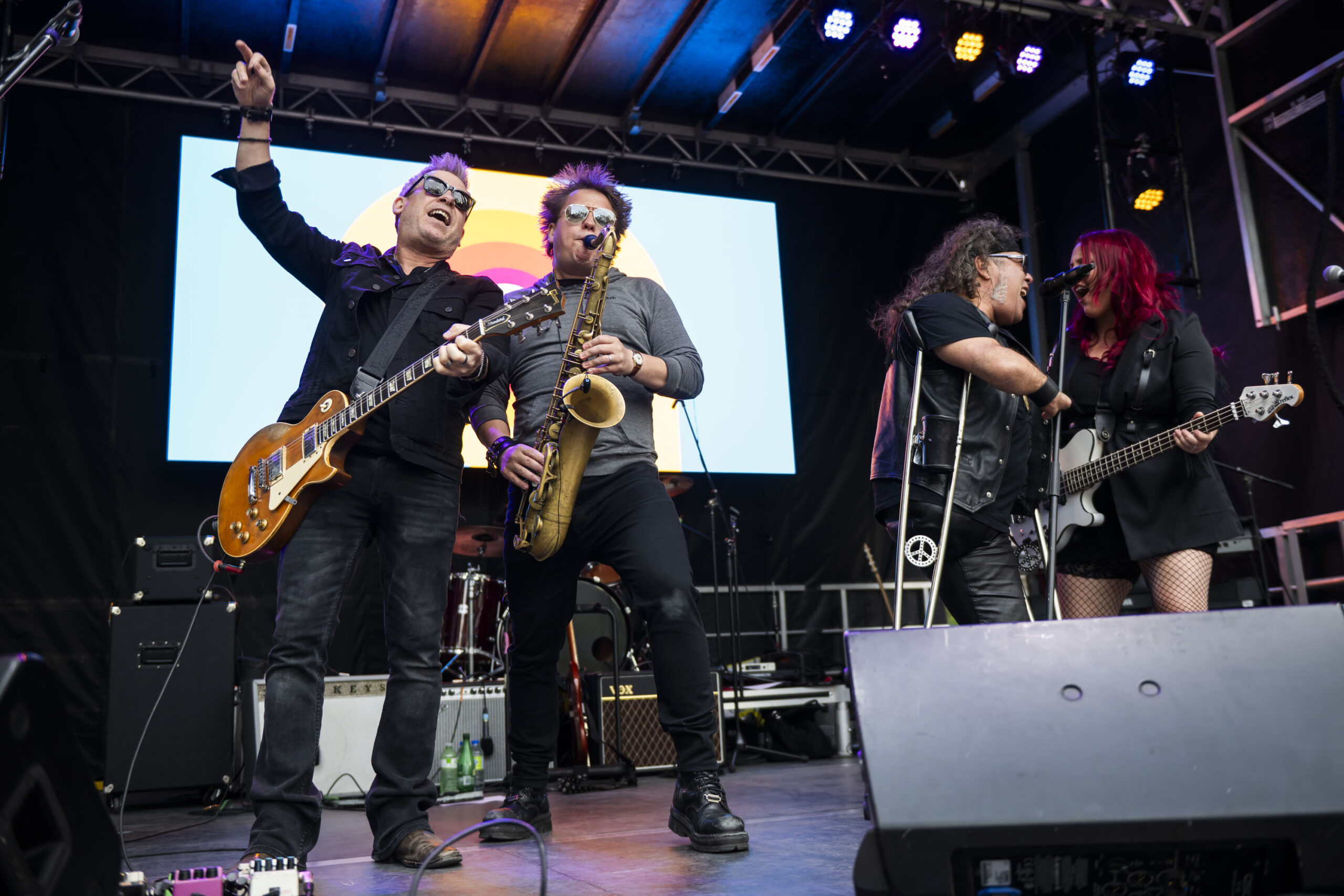 A lively band performs on an outdoor stage. A guitarist plays energetically beside a saxophonist, while a vocalist with crutches and a bassist with bright red hair sing together at a microphone. Behind them, stage lights glow and a vibrant circular logo is displayed on a screen.