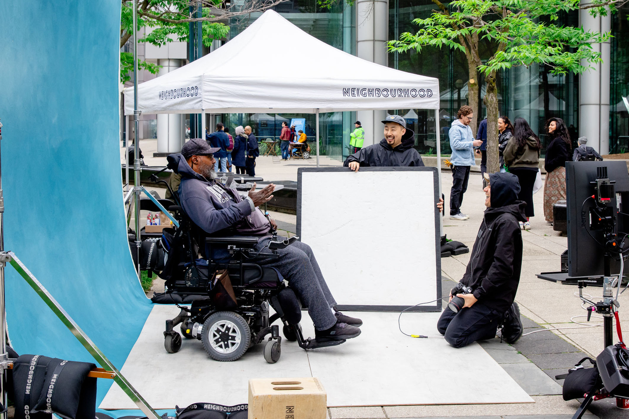 A smiling woman in a wheelchair interacts affectionately with a curly-haired service dog wearing a blue bandana and harness.