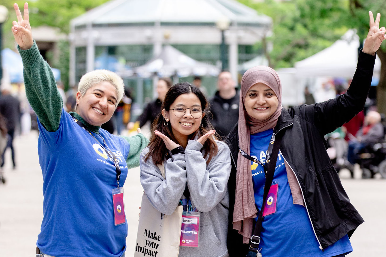 Three volunteers stand close together at AccessFest, smiling brightly and striking fun poses. The volunteer on the left, wearing a blue festival shirt and lanyard, raises both arms in peace signs.
