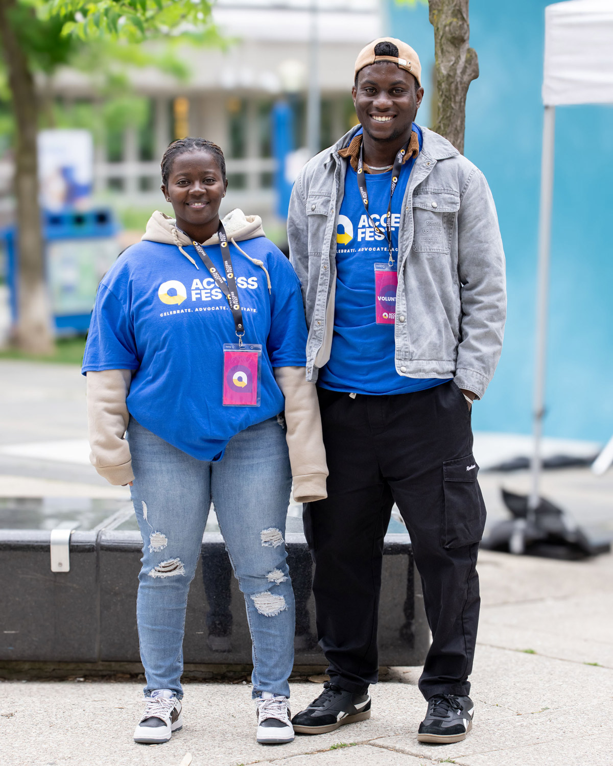 Two smiling AccessFest volunteers stand outdoors beside a tree. They are both wearing bright blue AccessFest t-shirts over their clothes, with purple volunteer badges around their necks.