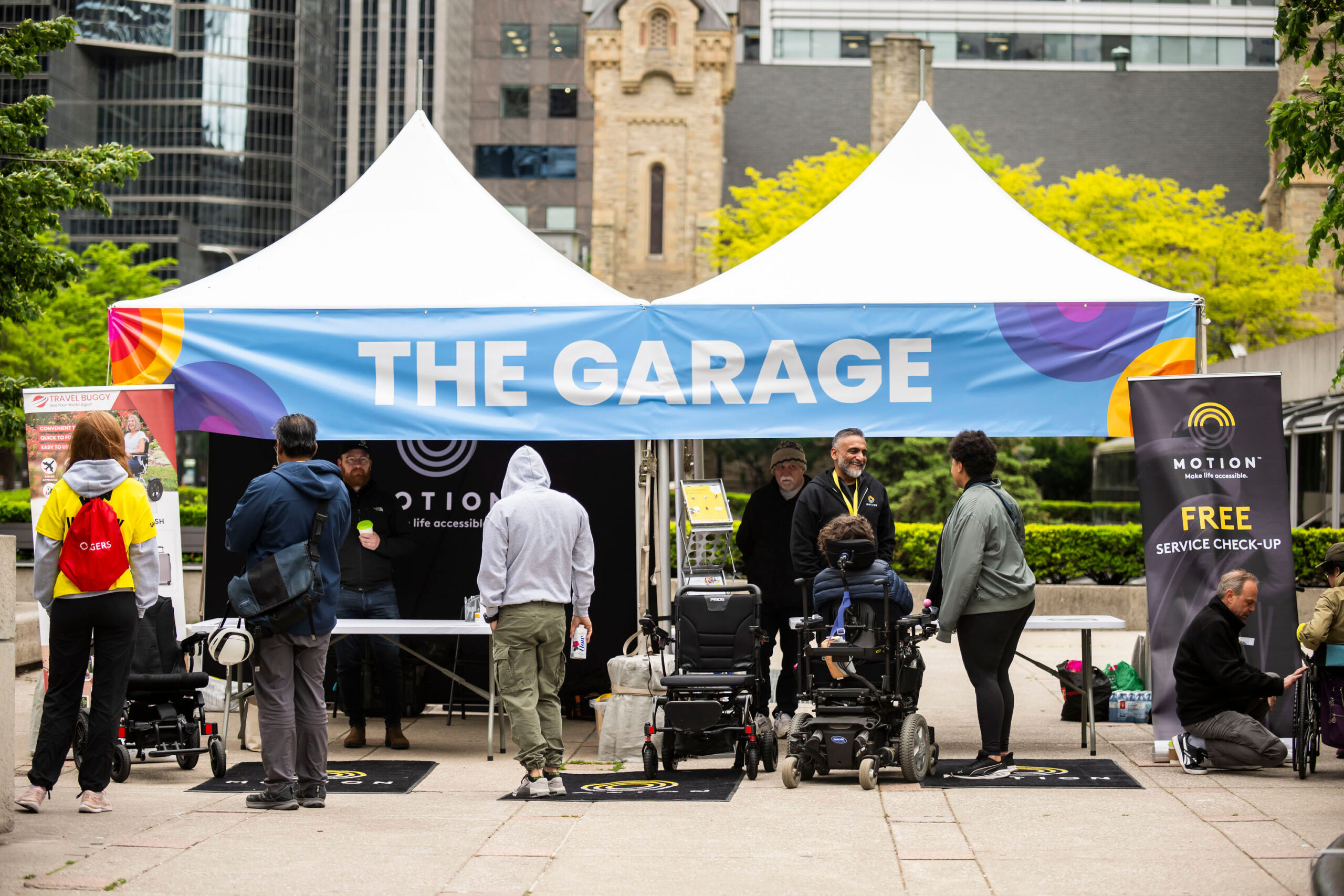 A group of people participate in a MapMission accessibility walk in an urban setting. One person in a wheelchair chats with another walking beside them, both smiling. Volunteers in blue vests hold green signs with white thumbs-up symbols. Tall buildings and a historic red train are visible in the background under a clear blue sky.
