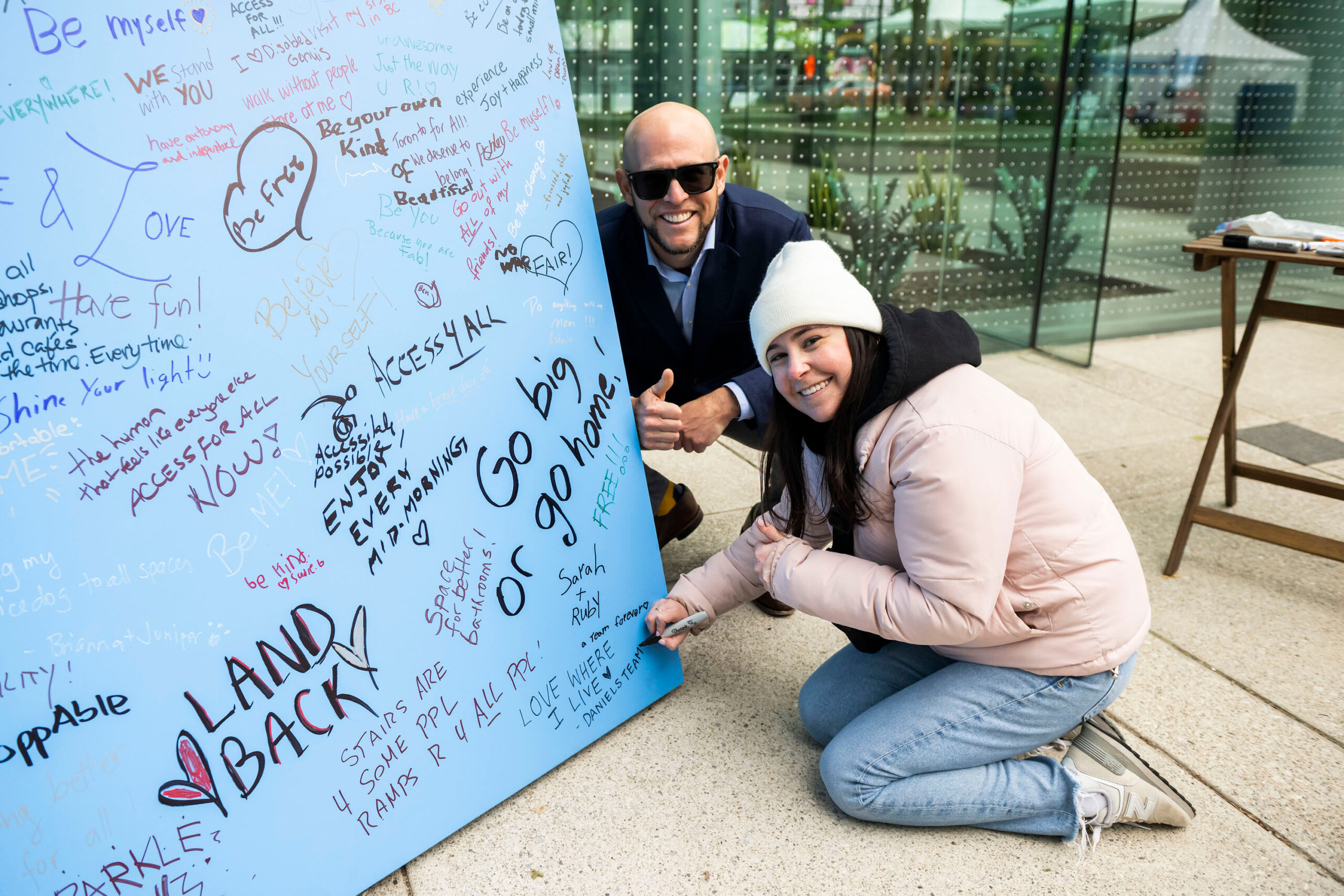 A large, inclusive outdoor yoga class in a sunny park, with participants of all abilities, including wheelchair users, stretching together in front of a lively crowd and white tents. The scene is framed by green trees and warm, golden light.