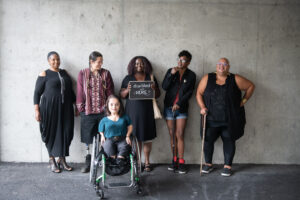 Six disabled people of color smile and pose in front of a concrete wall. Five people stand in the back, with a Black woman in the center holding up a chalkboard sign reading "disabled and here." A South Asian person in a wheelchair sits in front.