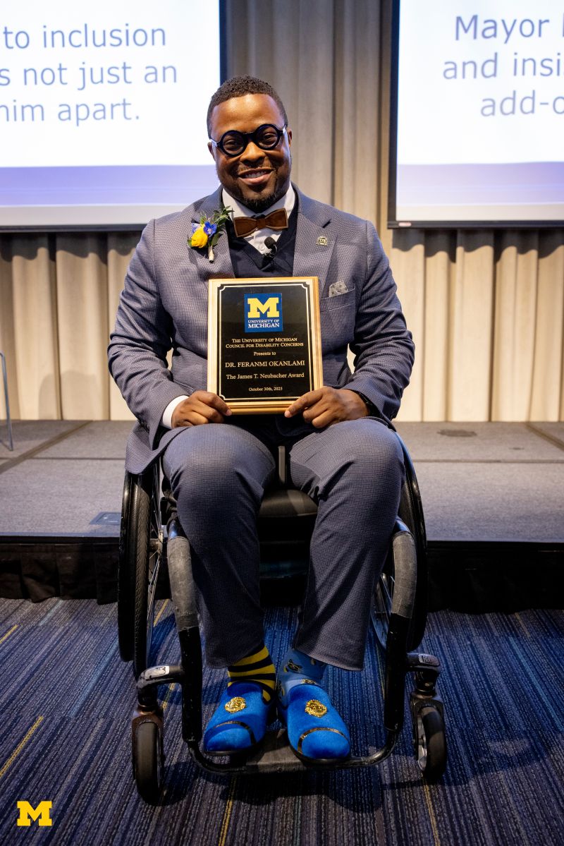 Feranmi Okanlami MD MS being named the 2025 recipient of U-M's James T. Neubacher Award. Seated in his wheelchair, he is smiling and holding his award.