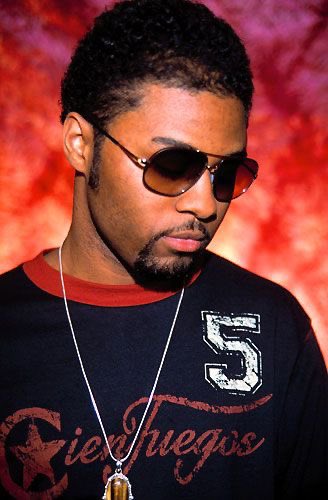 musiq soulchild wearing glasses, a chian, and looking down with a bright red background