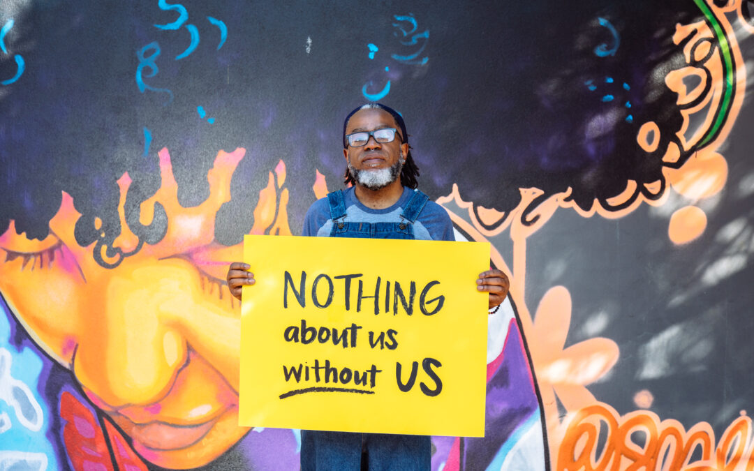 A Deaf Black man wearing glasses looks neutrally at the camera while holding a hand lettered sign declaring “NOTHING about us without US.” The man wears denim overalls and has a black and white beard, septum piercing, and hair pulled back with a bandana. The background features a vibrant mural of a Black woman looking down.