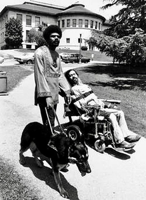 Image of two disabled men strolling and rolling down an outdoor pathway. Man on the left is Donald Galloway, tall Black man with an afro with a guide dog by his side. Man on the right is Ed Roberts, white man who is in a wheelchair. Both men are facing the direction of the camera while in mid-stroll/roll