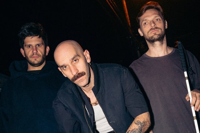 Three members of the band X Ambassadors pose together in a dimly lit setting. The central member, with a shaved head and mustache, leans forward toward the camera, while the two others stand slightly behind him. One member holds a white cane, highlighting disability representation within the group.